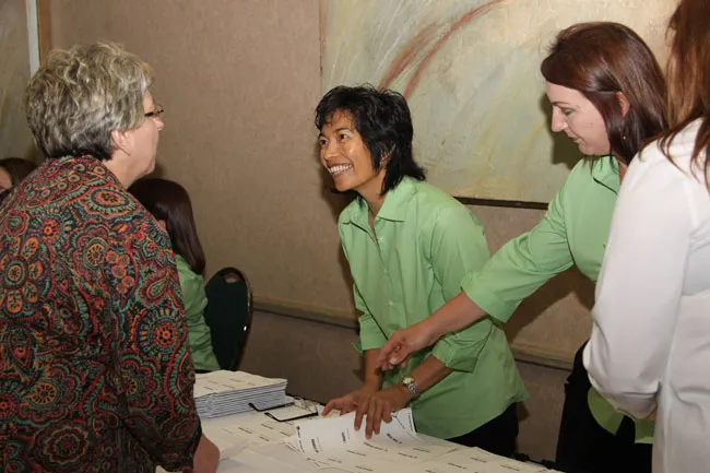 Group interaction: woman in green shirt handing documents to seated attendees at a table; collaborative setting