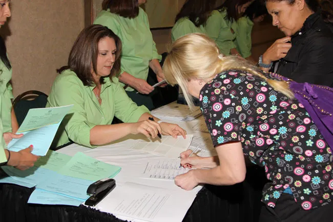 Two women reviewing paperwork at table—one in green shirt, one in floral top—with others nearby