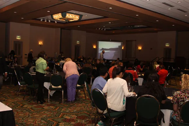 Large conference room with attendees seated at round tables, presenter on screen, ambient lighting