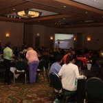 Large conference room with attendees seated at round tables, presenter on screen, ambient lighting