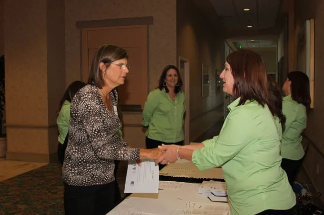 Two women shaking hands at registration table; others in green shirts observe in hallway setting