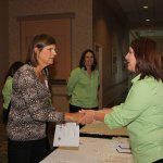 Two women shaking hands at registration table; others in green shirts observe in hallway setting