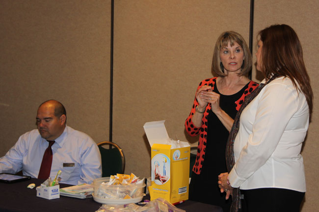 Two women interacting near table with yellow box and snacks; professional casual attire