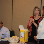 Two women interacting near table with yellow box and snacks; professional casual attire