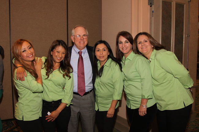 Group photo: man in suit with tie surrounded by five women in matching green shirts, smiling