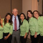Group photo: man in suit with tie surrounded by five women in matching green shirts, smiling