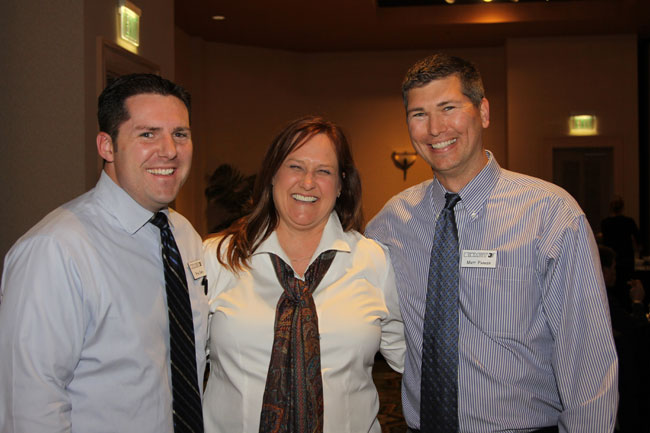 Three staff (two men, one woman) in light shirts and ties, smiling for group photo indoors