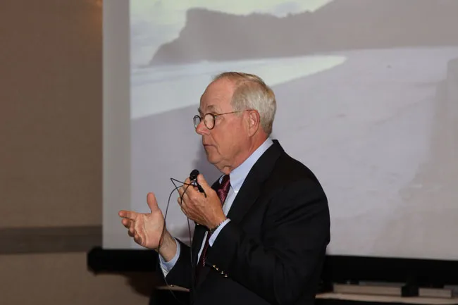Man in suit and tie gesturing while speaking at podium; projection screen visible behind him
