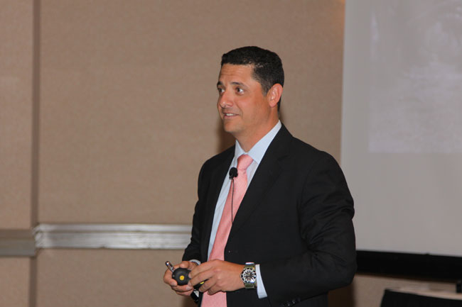 Man in dark suit and pink tie smiling, holding remote clicker; presentation screen in background