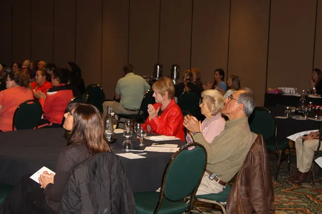 Audience seated at round tables during conference session; some taking notes, others listening