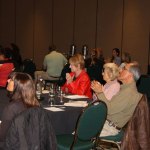 Audience seated at round tables during conference session; some taking notes, others listening