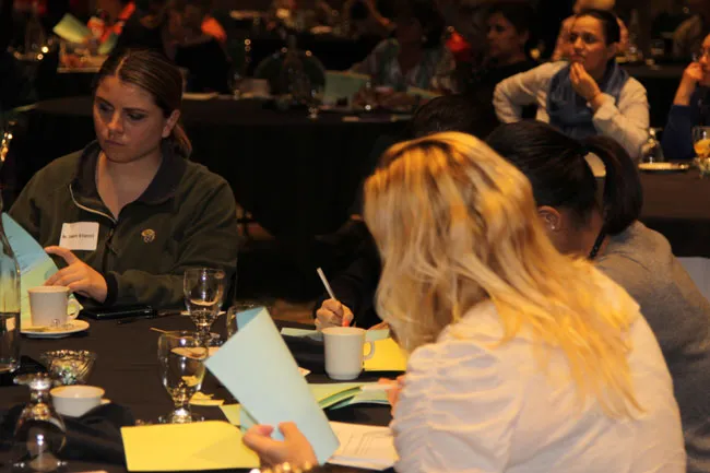 Close-up of attendees at table: woman writing, another reviewing materials, engaged in session