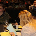 Close-up of attendees at table: woman writing, another reviewing materials, engaged in session