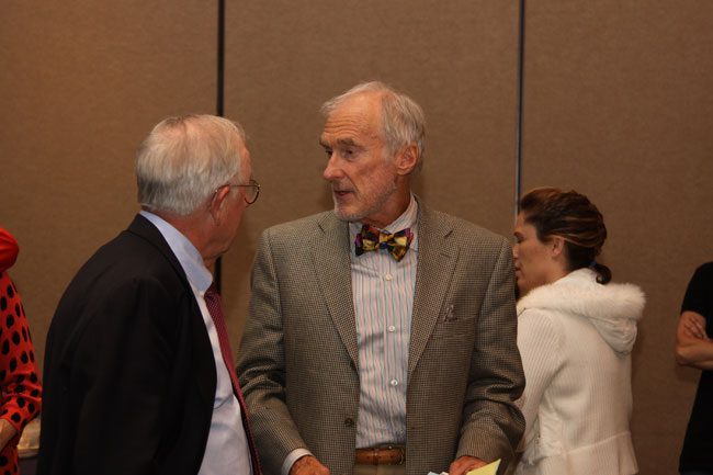 Two men in formal attire conversing indoors: one in dark suit with red tie, the other in gray suit with colorful bow tie