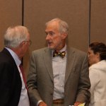 Two men in formal attire conversing indoors: one in dark suit with red tie, the other in gray suit with colorful bow tie